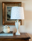 White lamp with blue patterned shade on a wooden table next to a framed picture