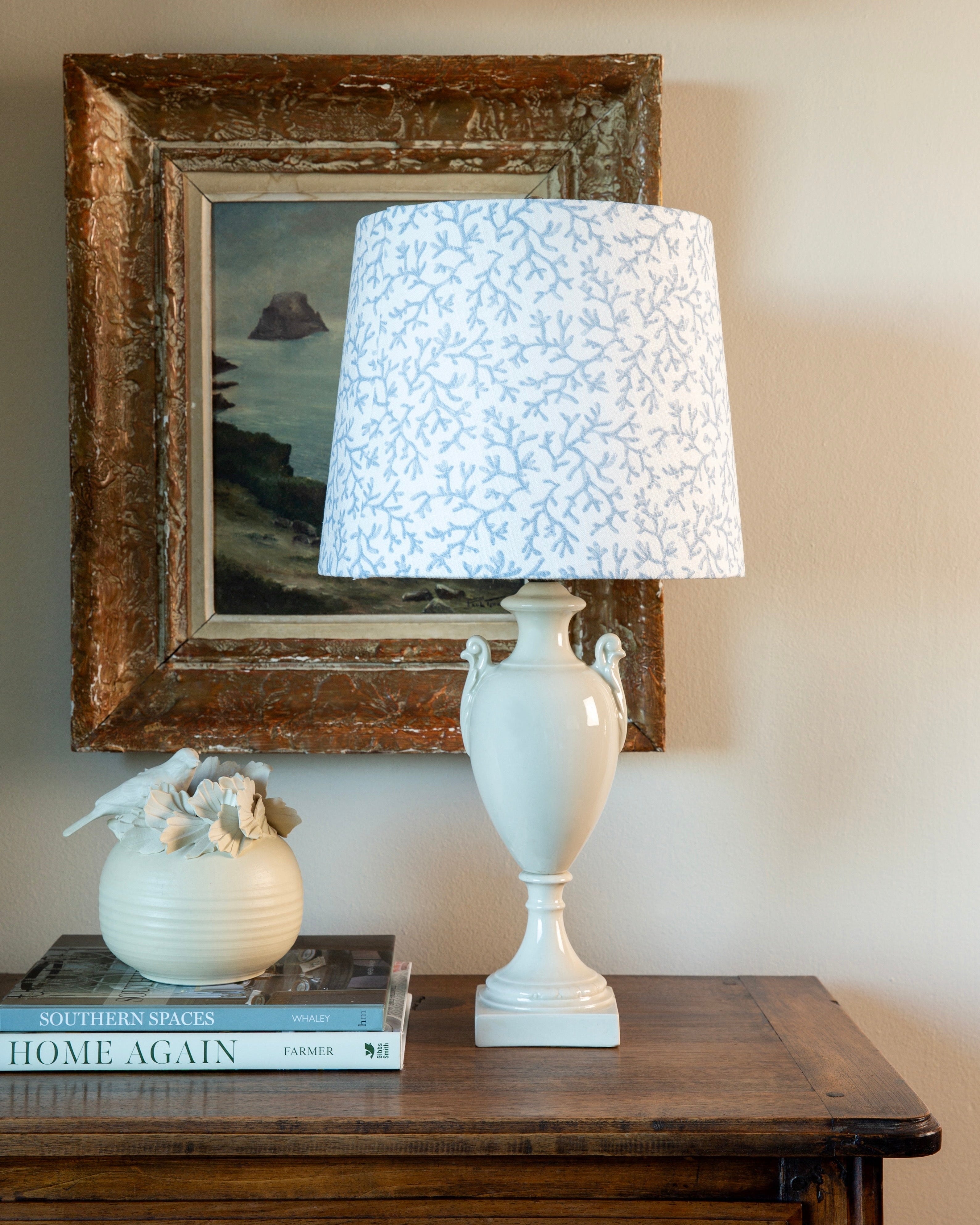 White lamp with blue patterned shade on a wooden table next to a framed picture